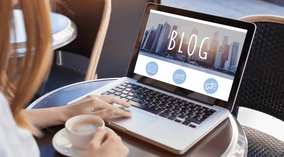 ノートパソコンの画面に「BLOG」と書かれたブログサイトの画面が表示されており、カフェでコーヒーを飲みながら作業している様子の写真。